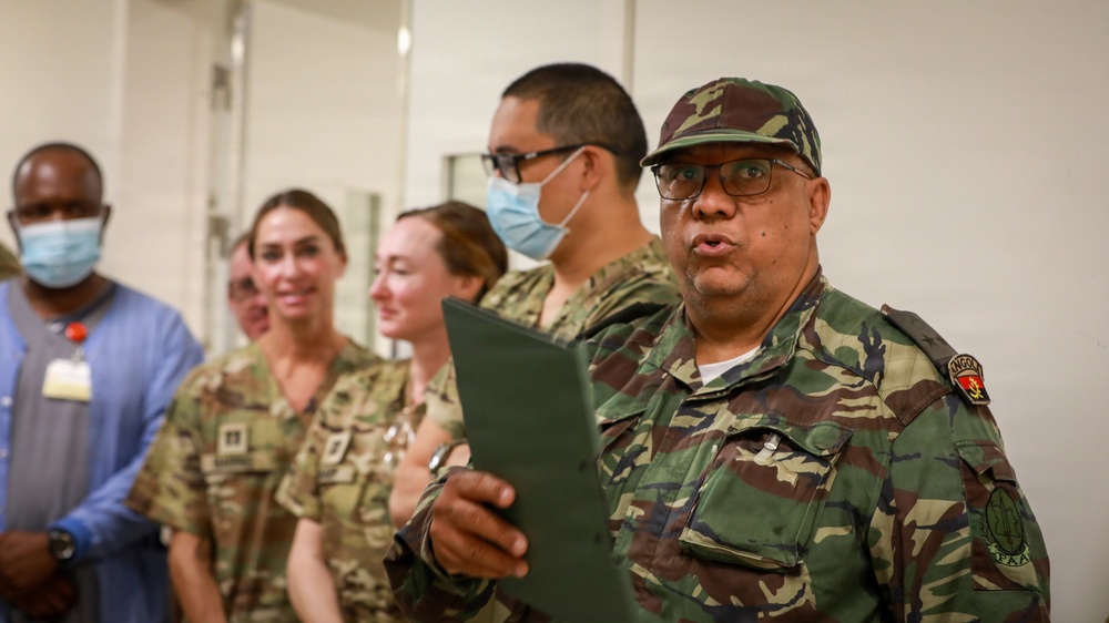 A group of military personnel and civilians are gathered in what appears to be an indoor facility. The foreground figure, wearing a camouflage uniform with a visible shoulder patch reading 'ANGOLA', is holding a dark folder and appears to be speaking. Several other individuals in military uniforms and civilian attire are visible in the background, some wearing blue surgical masks.