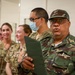 A group of military personnel and civilians are gathered in what appears to be an indoor facility. The foreground figure, wearing a camouflage uniform with a visible shoulder patch reading 'ANGOLA', is holding a dark folder and appears to be speaking. Several other individuals in military uniforms and civilian attire are visible in the background, some wearing blue surgical masks.