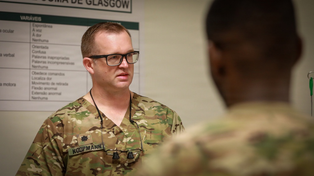 A U.S. military medical officer wearing OCP (Operational Camouflage Pattern) uniform with the name tape 'KOOPMANN' and a Major rank insignia is engaged in conversation with another service member. They appear to be in a medical training or clinical environment, with a Portuguese-language Glasgow Coma Scale chart visible on the wall behind them. The setting suggests a joint military medical training exercise or international military cooperation event.
