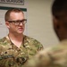 A U.S. military medical officer wearing OCP (Operational Camouflage Pattern) uniform with the name tape 'KOOPMANN' and a Major rank insignia is engaged in conversation with another service member. They appear to be in a medical training or clinical environment, with a Portuguese-language Glasgow Coma Scale chart visible on the wall behind them. The setting suggests a joint military medical training exercise or international military cooperation event.