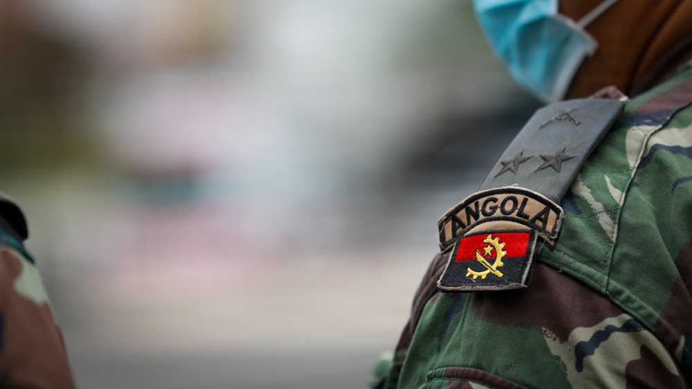 The image shows a close-up of a military uniform shoulder area belonging to an Angolan armed forces member. A patch displaying the Angolan flag and the text 'ANGOLA' is clearly visible on the upper arm, along with rank insignia showing four stars on an epaulette. The person is also wearing a light blue surgical/medical face mask, visible at the top of the frame.