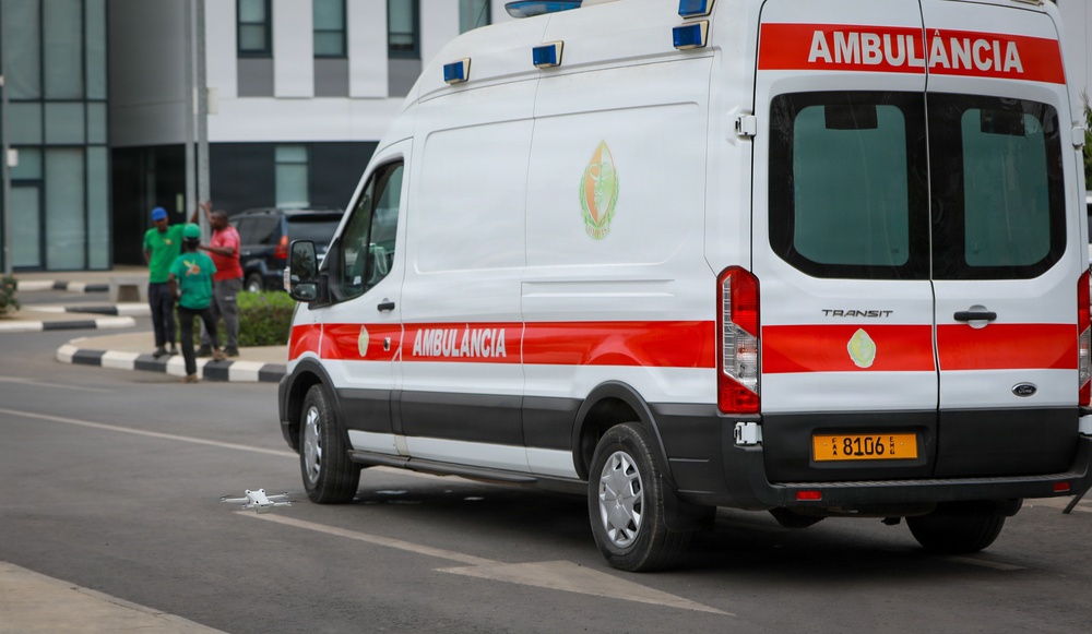 A white and red Ford Transit ambulance labeled 'AMBULÀNCIA' is parked or slowly moving on a road in front of a modern building. The vehicle bears a yellow license plate reading 'CAA 8106 EA' and displays a green circular emblem, suggesting a Portuguese-speaking African country, likely Angola or Mozambique. Several people in casual clothing are visible in the background near a median strip.
