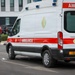 A white and red Ford Transit ambulance labeled 'AMBULÀNCIA' is parked or slowly moving on a road in front of a modern building. The vehicle bears a yellow license plate reading 'CAA 8106 EA' and displays a green circular emblem, suggesting a Portuguese-speaking African country, likely Angola or Mozambique. Several people in casual clothing are visible in the background near a median strip.