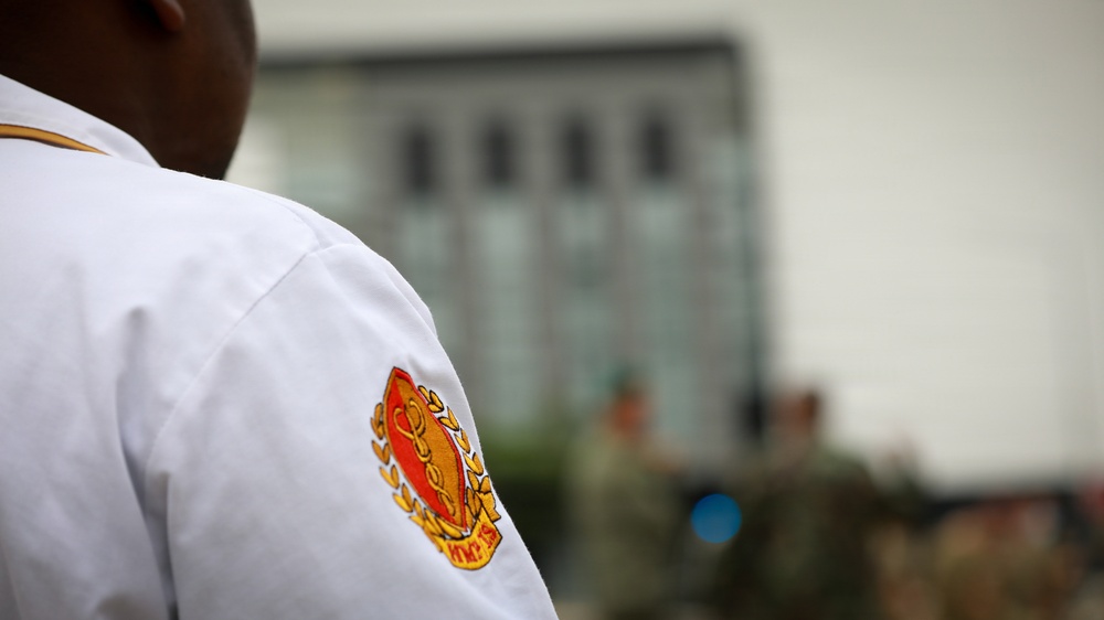 A close-up photograph focusing on the shoulder of a person wearing a white uniform with an embroidered patch or insignia in red, orange, and gold. The patch appears to feature a crest or shield design with laurel wreath elements. The background is blurred, showing what appears to be a building with large windows and other individuals in the distance.