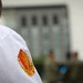 A close-up photograph focusing on the shoulder of a person wearing a white uniform with an embroidered patch or insignia in red, orange, and gold. The patch appears to feature a crest or shield design with laurel wreath elements. The background is blurred, showing what appears to be a building with large windows and other individuals in the distance.