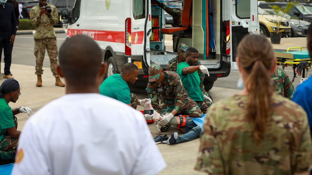 The image depicts what appears to be a military medical training exercise or mass casualty drill conducted outdoors near an ambulance. Personnel in camouflage uniforms and green medical scrubs are providing simulated treatment to individuals on the ground. Observers in military uniforms, including at least one person photographing the exercise, are watching from the periphery.