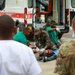 The image depicts what appears to be a military medical training exercise or mass casualty drill conducted outdoors near an ambulance. Personnel in camouflage uniforms and green medical scrubs are providing simulated treatment to individuals on the ground. Observers in military uniforms, including at least one person photographing the exercise, are watching from the periphery.