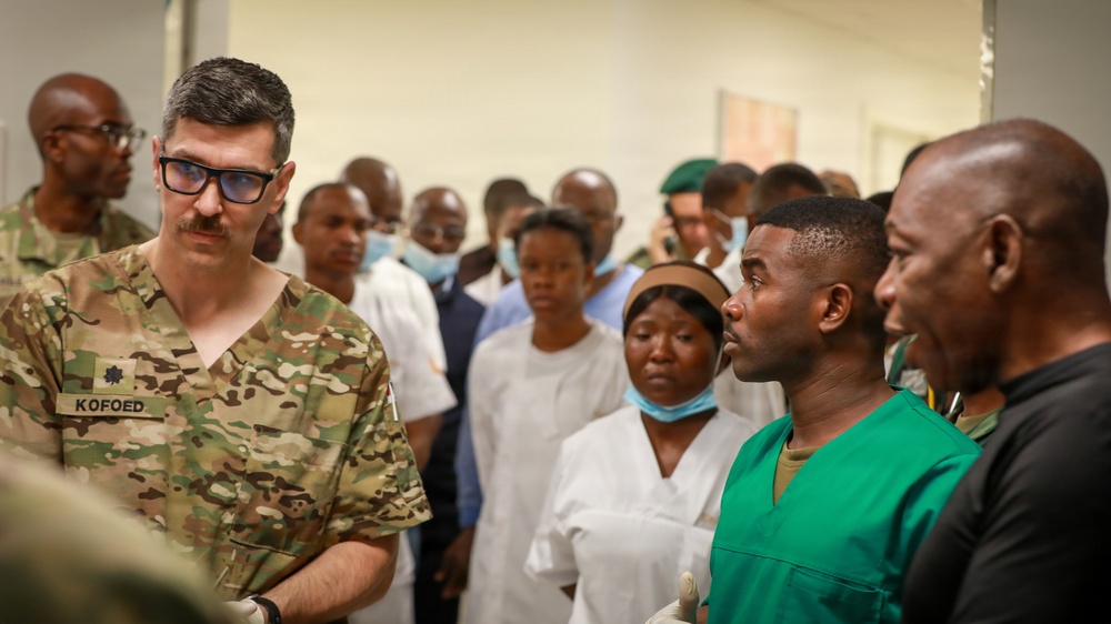 A military medical professional identified by the name tape 'KOFOED' wearing U.S. Army OCP (Operational Camouflage Pattern) uniform is engaged in conversation with local medical staff in what appears to be a medical facility. The scene suggests a military medical training, advisory, or humanitarian assistance mission, with a large group of local healthcare workers gathered in the background. The interaction appears to be a briefing or consultation between U.S. military medical personnel and host-nation medical staff.