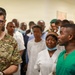A military medical professional identified by the name tape 'KOFOED' wearing U.S. Army OCP (Operational Camouflage Pattern) uniform is engaged in conversation with local medical staff in what appears to be a medical facility. The scene suggests a military medical training, advisory, or humanitarian assistance mission, with a large group of local healthcare workers gathered in the background. The interaction appears to be a briefing or consultation between U.S. military medical personnel and host-nation medical staff.