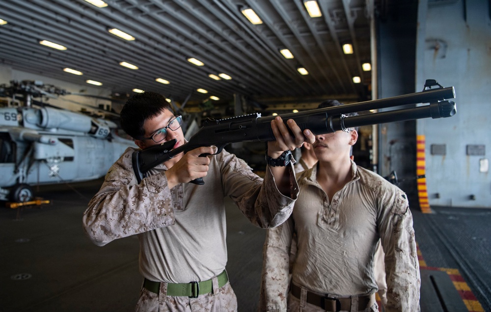 Two military personnel in desert camouflage uniforms are handling a shotgun inside what appears to be a naval vessel's hangar bay. One individual is aiming the weapon while the other assists or observes closely. A military helicopter marked with the number '69' is visible in the background.