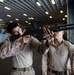 Two military personnel in desert camouflage uniforms are handling a shotgun inside what appears to be a naval vessel's hangar bay. One individual is aiming the weapon while the other assists or observes closely. A military helicopter marked with the number '69' is visible in the background.