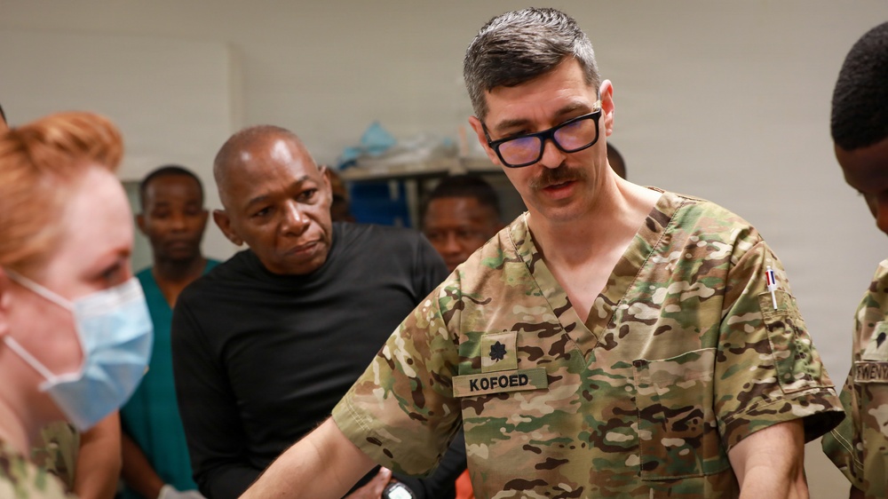 A U.S. military medical personnel identified as 'KOFOED' wearing camouflage scrubs and rank insignia of a Major (oak leaf cluster) is engaged in what appears to be a medical training or demonstration session. He is surrounded by a mixed group of military and civilian personnel, including a colleague in scrubs with a surgical mask. The setting appears to be a medical facility or clinical training environment.