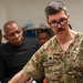 A U.S. military medical personnel identified as 'KOFOED' wearing camouflage scrubs and rank insignia of a Major (oak leaf cluster) is engaged in what appears to be a medical training or demonstration session. He is surrounded by a mixed group of military and civilian personnel, including a colleague in scrubs with a surgical mask. The setting appears to be a medical facility or clinical training environment.
