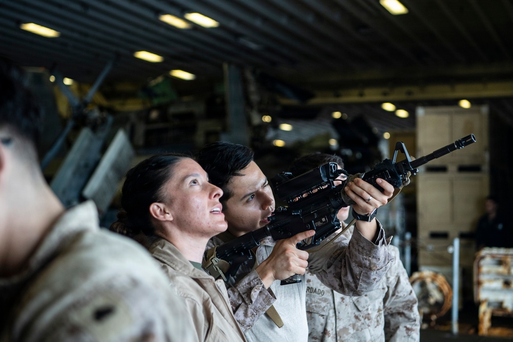 Two military personnel in desert camouflage uniforms are engaged in what appears to be a weapons handling or training exercise inside a large enclosed military facility, likely a ship's hangar deck or similar structure. One service member is aiming an M16/M4-style rifle upward while another observes and appears to be providing instruction or guidance. The setting includes overhead lighting, military equipment, and storage areas visible in the background.