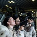 Two military personnel in desert camouflage uniforms are engaged in what appears to be a weapons handling or training exercise inside a large enclosed military facility, likely a ship's hangar deck or similar structure. One service member is aiming an M16/M4-style rifle upward while another observes and appears to be providing instruction or guidance. The setting includes overhead lighting, military equipment, and storage areas visible in the background.