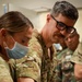 Military medical personnel in OCP (Operational Camouflage Pattern) uniforms are performing what appears to be a medical training exercise or patient care procedure. At least three individuals in camouflage uniforms are leaning over what appears to be a medical mannequin or patient, with one person wearing a surgical mask and another wearing black-framed glasses. A name tape reading 'KOPUS' (or similar) is partially visible on one uniform.