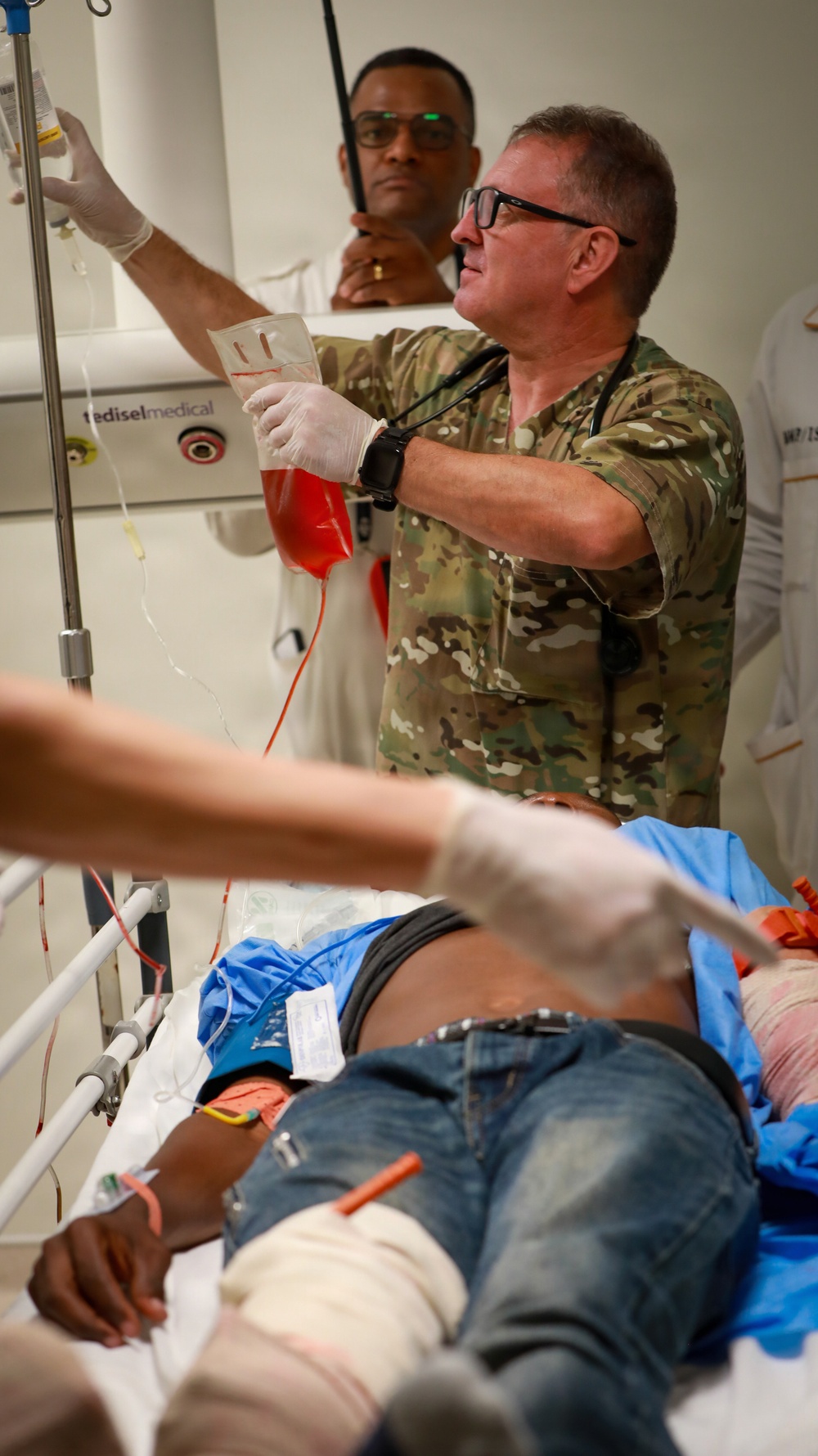 A military medical personnel in camouflage uniform is administering a blood transfusion to a patient lying on a medical bed. At least two medical providers wearing gloves are actively treating the patient, who has visible bandaging and multiple IV lines. The setting appears to be a medical treatment facility or trauma bay.