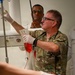 A military medical personnel in camouflage uniform is administering a blood transfusion to a patient lying on a medical bed. At least two medical providers wearing gloves are actively treating the patient, who has visible bandaging and multiple IV lines. The setting appears to be a medical treatment facility or trauma bay.