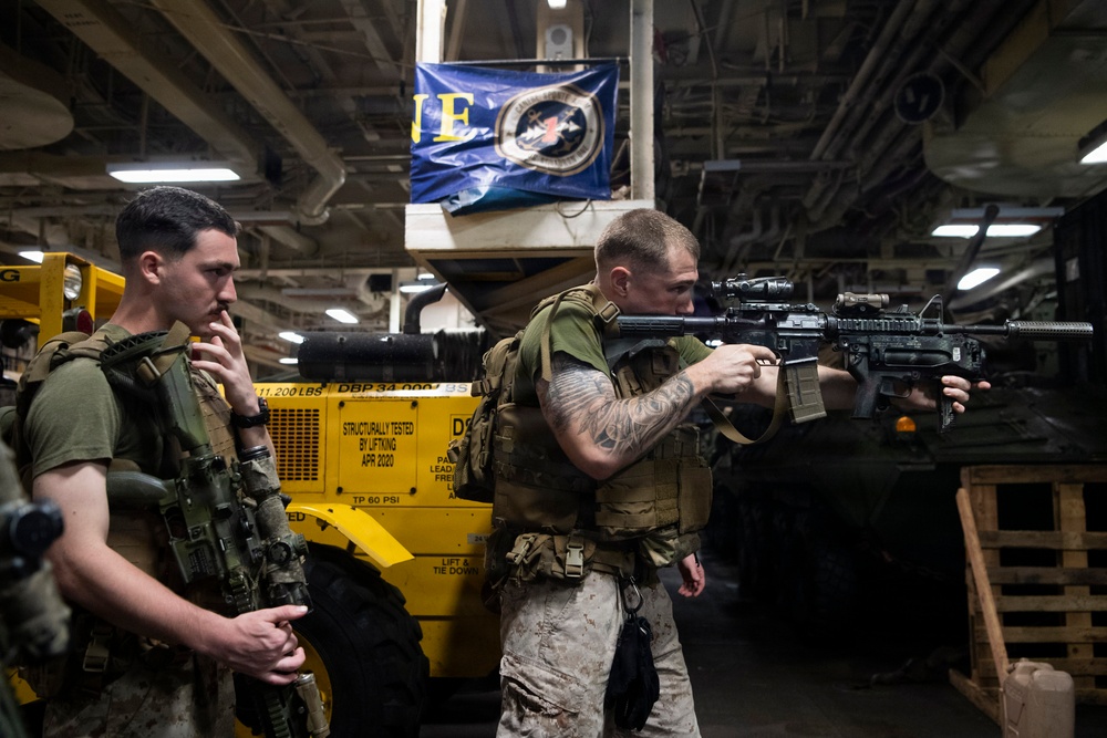 Two armed military personnel, appearing to be U.S. Marines based on their camouflage uniforms and gear, are positioned inside what appears to be a ship's hangar or cargo deck. One Marine is aiming an M4-style carbine rifle while the other stands nearby in a tactical posture. The setting suggests a training exercise or security drill aboard a naval vessel.