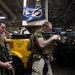Two armed military personnel, appearing to be U.S. Marines based on their camouflage uniforms and gear, are positioned inside what appears to be a ship's hangar or cargo deck. One Marine is aiming an M4-style carbine rifle while the other stands nearby in a tactical posture. The setting suggests a training exercise or security drill aboard a naval vessel.