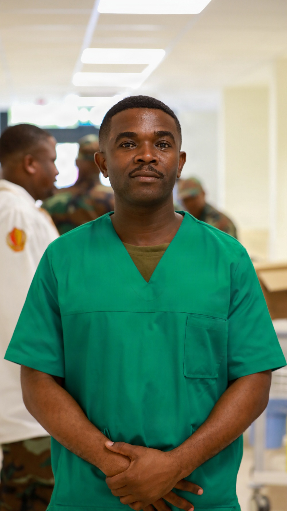 A man wearing green medical scrubs stands in the foreground of what appears to be a medical or clinical facility. He has a composed expression with hands clasped in front of him. In the background, several individuals in military camouflage uniforms are visible, suggesting a military medical setting.