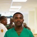 A man wearing green medical scrubs stands in the foreground of what appears to be a medical or clinical facility. He has a composed expression with hands clasped in front of him. In the background, several individuals in military camouflage uniforms are visible, suggesting a military medical setting.