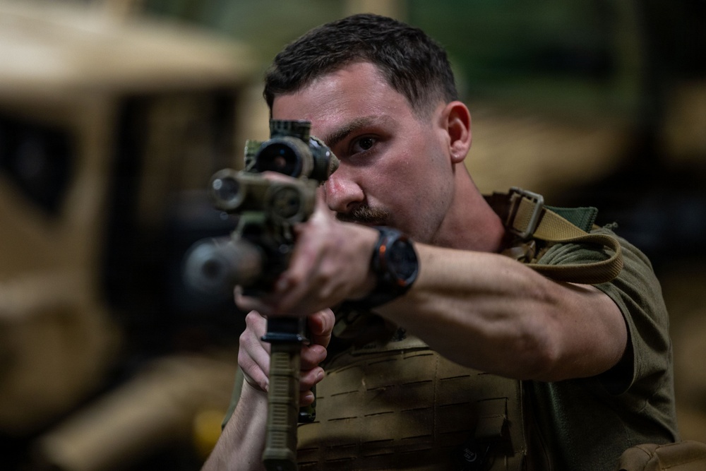 A male soldier in military gear is aiming a rifle with an optical scope directly toward the camera in what appears to be an indoor military facility or motor pool. He is wearing a plate carrier vest, olive drab uniform, and a tactical watch. Military vehicles are visible in the blurred background.