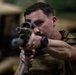 A male soldier in military gear is aiming a rifle with an optical scope directly toward the camera in what appears to be an indoor military facility or motor pool. He is wearing a plate carrier vest, olive drab uniform, and a tactical watch. Military vehicles are visible in the blurred background.