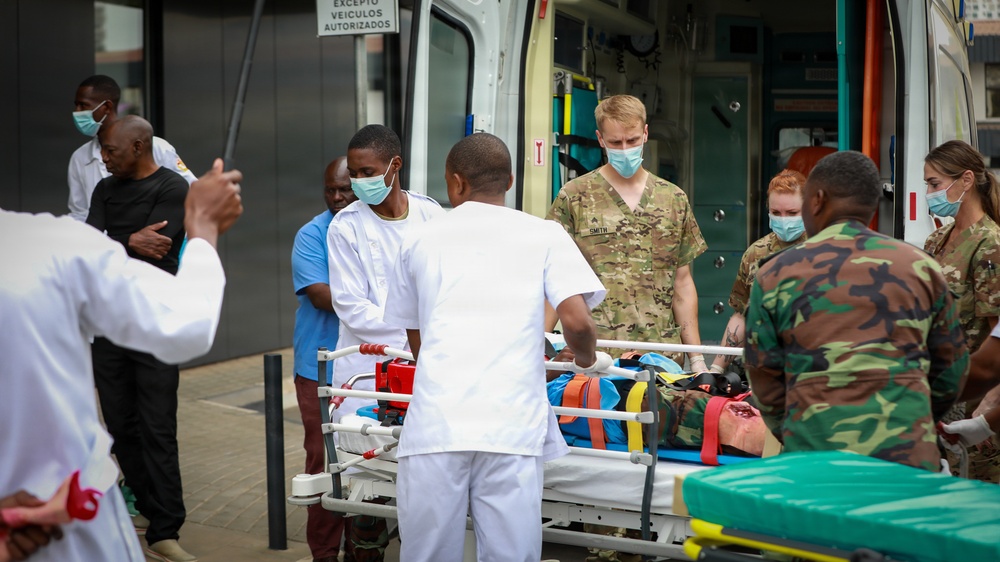 A medical emergency scene outside what appears to be a hospital or medical facility, showing a collaborative effort between military personnel and civilian medical staff. Multiple individuals in various uniforms — including military camouflage, scrubs, and white medical attire — are gathered around a stretcher being unloaded from an ambulance. A sign in Portuguese ('Excepto Veículos Autorizados') suggests the location is in a Portuguese-speaking country.