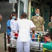 A medical emergency scene outside what appears to be a hospital or medical facility, showing a collaborative effort between military personnel and civilian medical staff. Multiple individuals in various uniforms — including military camouflage, scrubs, and white medical attire — are gathered around a stretcher being unloaded from an ambulance. A sign in Portuguese ('Excepto Veículos Autorizados') suggests the location is in a Portuguese-speaking country.