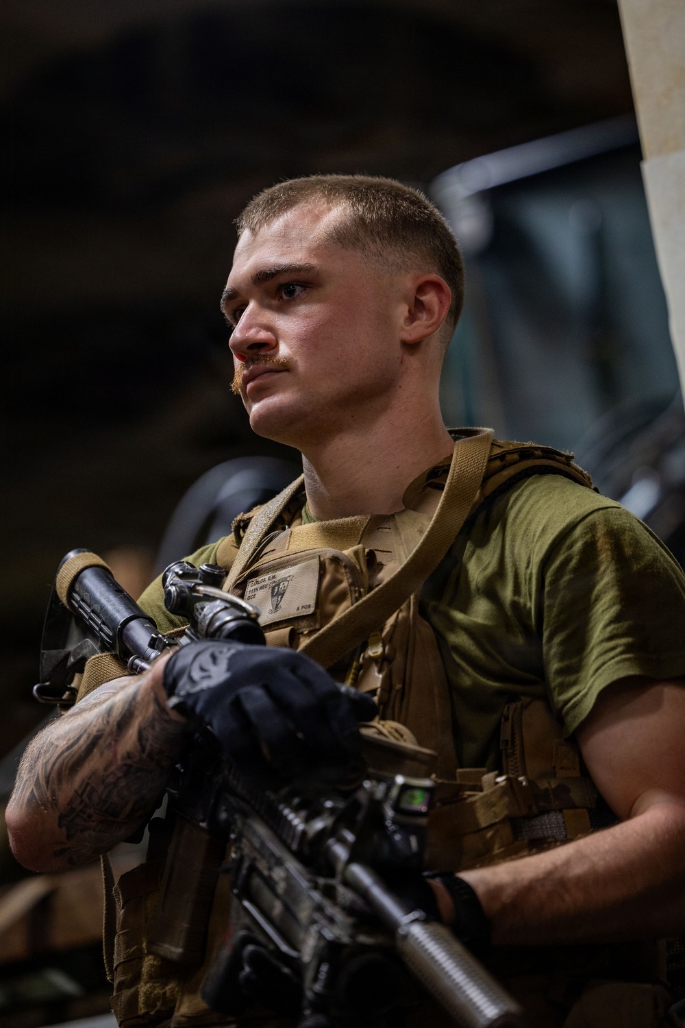 A young male service member in tactical gear holds a rifle while looking off to the side. He is wearing a plate carrier vest over an olive green t-shirt, black tactical gloves, and has visible tattoos on his forearm. A name tape and unit patch are visible on his vest.