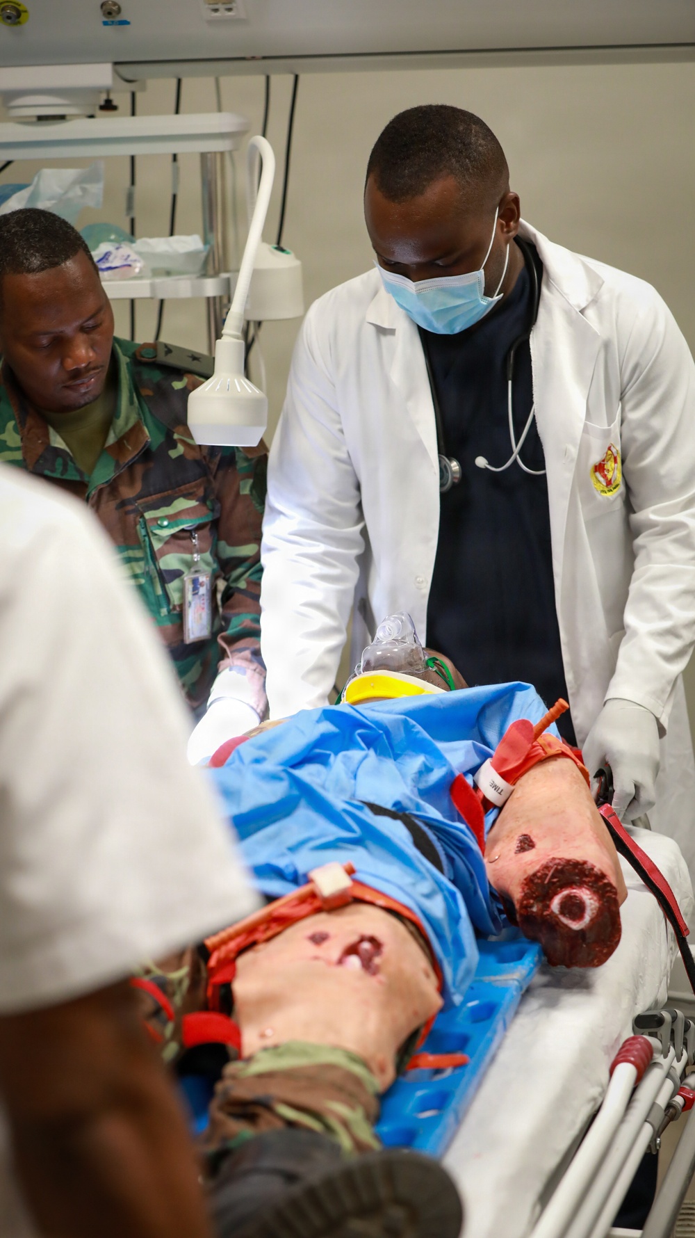 A medical training or simulation exercise is underway in a clinical setting. A physician in a white lab coat and surgical mask, along with a person in military camouflage uniform, are attending to a medical training manikin on a gurney. The manikin displays simulated traumatic injuries including a wound on the arm with a tourniquet applied, consistent with combat casualty care training.