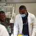 A medical training or simulation exercise is underway in a clinical setting. A physician in a white lab coat and surgical mask, along with a person in military camouflage uniform, are attending to a medical training manikin on a gurney. The manikin displays simulated traumatic injuries including a wound on the arm with a tourniquet applied, consistent with combat casualty care training.