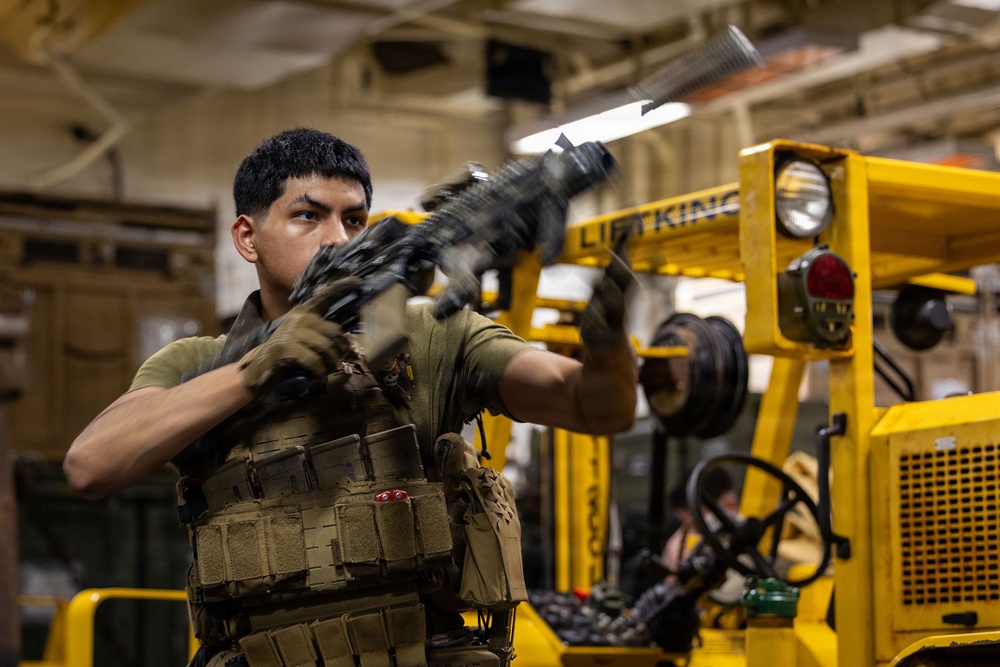 A military service member wearing tactical gear and a plate carrier vest is handling a rifle in what appears to be a ship's hangar bay or cargo deck. The individual is inspecting or clearing the weapon while standing near yellow industrial equipment. The setting suggests a naval vessel based on the overhead infrastructure and yellow forklift equipment visible in the background.