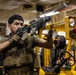 A military service member wearing tactical gear and a plate carrier vest is handling a rifle in what appears to be a ship's hangar bay or cargo deck. The individual is inspecting or clearing the weapon while standing near yellow industrial equipment. The setting suggests a naval vessel based on the overhead infrastructure and yellow forklift equipment visible in the background.