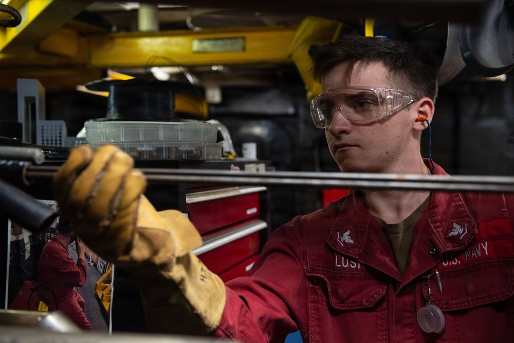 A U.S. Navy sailor wearing a red uniform, safety goggles, and leather work gloves is performing maintenance or mechanical work in what appears to be a shipboard workshop or machinery space. The sailor's name tag reads 'LUST' and the uniform is marked 'U.S. NAVY'. The environment is an industrial workspace with tool chests, equipment, and overhead structures visible.