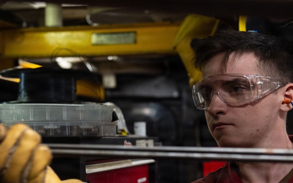 A U.S. Navy sailor wearing a red uniform, safety goggles, and leather work gloves is performing maintenance or mechanical work in what appears to be a shipboard workshop or machinery space. The sailor's name tag reads 'LUST' and the uniform is marked 'U.S. NAVY'. The environment is an industrial workspace with tool chests, equipment, and overhead structures visible.