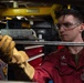 A U.S. Navy sailor wearing a red uniform, safety goggles, and leather work gloves is performing maintenance or mechanical work in what appears to be a shipboard workshop or machinery space. The sailor's name tag reads 'LUST' and the uniform is marked 'U.S. NAVY'. The environment is an industrial workspace with tool chests, equipment, and overhead structures visible.