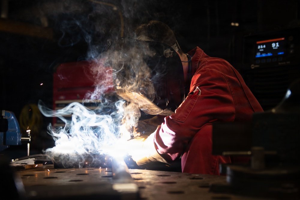 A welder in red protective coveralls and a Miller welding helmet is actively welding on a metal workbench in an industrial workshop. Bright arc light, sparks, and billowing smoke/fumes are visible at the weld point. A welding machine with a digital display is visible in the background.