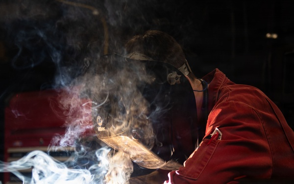 A welder in red protective coveralls and a Miller welding helmet is actively welding on a metal workbench in an industrial workshop. Bright arc light, sparks, and billowing smoke/fumes are visible at the weld point. A welding machine with a digital display is visible in the background.