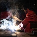 A welder in red protective coveralls and a Miller welding helmet is actively welding on a metal workbench in an industrial workshop. Bright arc light, sparks, and billowing smoke/fumes are visible at the weld point. A welding machine with a digital display is visible in the background.