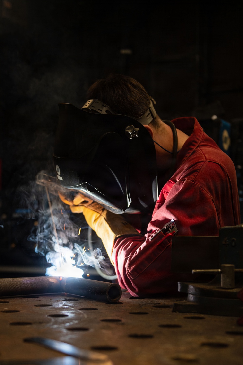 A welder is actively welding a metal pipe or rod onto a perforated steel work table in a dark industrial workshop. The welder is wearing a Miller brand auto-darkening welding helmet, red coveralls, and heavy leather welding gloves. Bright welding arc light, sparks, and smoke are clearly visible at the point of contact.