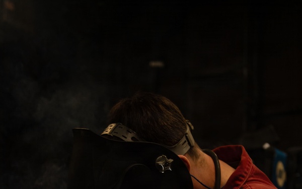 A welder is actively welding a metal pipe or rod onto a perforated steel work table in a dark industrial workshop. The welder is wearing a Miller brand auto-darkening welding helmet, red coveralls, and heavy leather welding gloves. Bright welding arc light, sparks, and smoke are clearly visible at the point of contact.