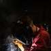 A welder is actively welding a metal pipe or rod onto a perforated steel work table in a dark industrial workshop. The welder is wearing a Miller brand auto-darkening welding helmet, red coveralls, and heavy leather welding gloves. Bright welding arc light, sparks, and smoke are clearly visible at the point of contact.