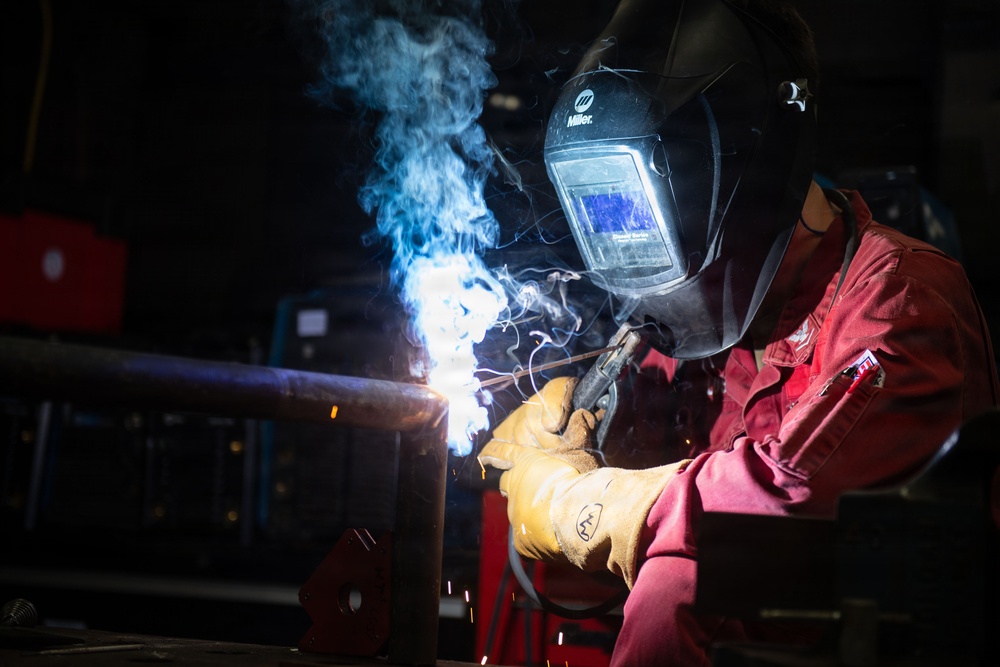 A welder is actively welding a metal pipe or structural component in an industrial workshop setting. The worker is wearing full protective gear including a Miller brand auto-darkening welding helmet and leather welding gloves, dressed in a red work uniform. Blue welding arc light, smoke, and sparks are visibly emanating from the weld point.
