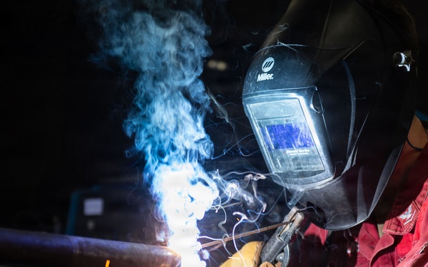 A welder is actively welding a metal pipe or structural component in an industrial workshop setting. The worker is wearing full protective gear including a Miller brand auto-darkening welding helmet and leather welding gloves, dressed in a red work uniform. Blue welding arc light, smoke, and sparks are visibly emanating from the weld point.