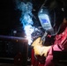 A welder is actively welding a metal pipe or structural component in an industrial workshop setting. The worker is wearing full protective gear including a Miller brand auto-darkening welding helmet and leather welding gloves, dressed in a red work uniform. Blue welding arc light, smoke, and sparks are visibly emanating from the weld point.