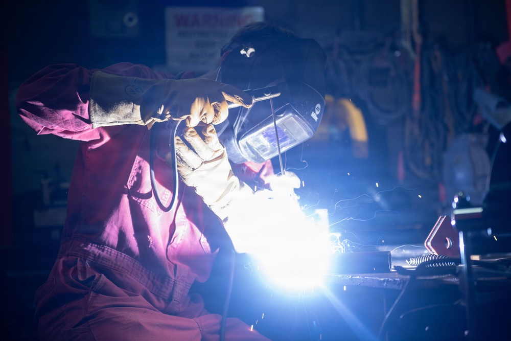 A welder is actively performing a welding operation in what appears to be an industrial workshop or fabrication facility. The worker is equipped with proper personal protective equipment including a welding helmet and heavy-duty gloves. The bright arc flash from the welding process illuminates the dark workspace with intense blue-white light and visible sparks.