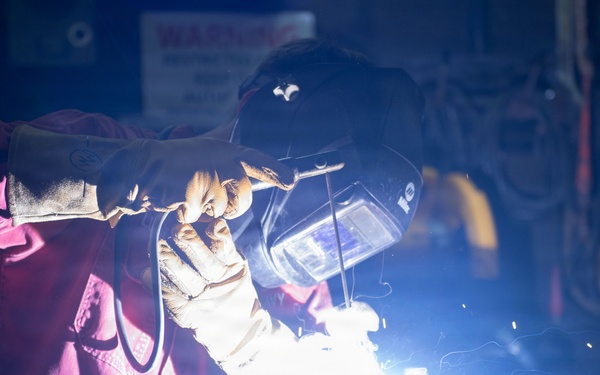 A welder is actively performing a welding operation in what appears to be an industrial workshop or fabrication facility. The worker is equipped with proper personal protective equipment including a welding helmet and heavy-duty gloves. The bright arc flash from the welding process illuminates the dark workspace with intense blue-white light and visible sparks.