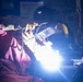 A welder is actively performing a welding operation in what appears to be an industrial workshop or fabrication facility. The worker is equipped with proper personal protective equipment including a welding helmet and heavy-duty gloves. The bright arc flash from the welding process illuminates the dark workspace with intense blue-white light and visible sparks.